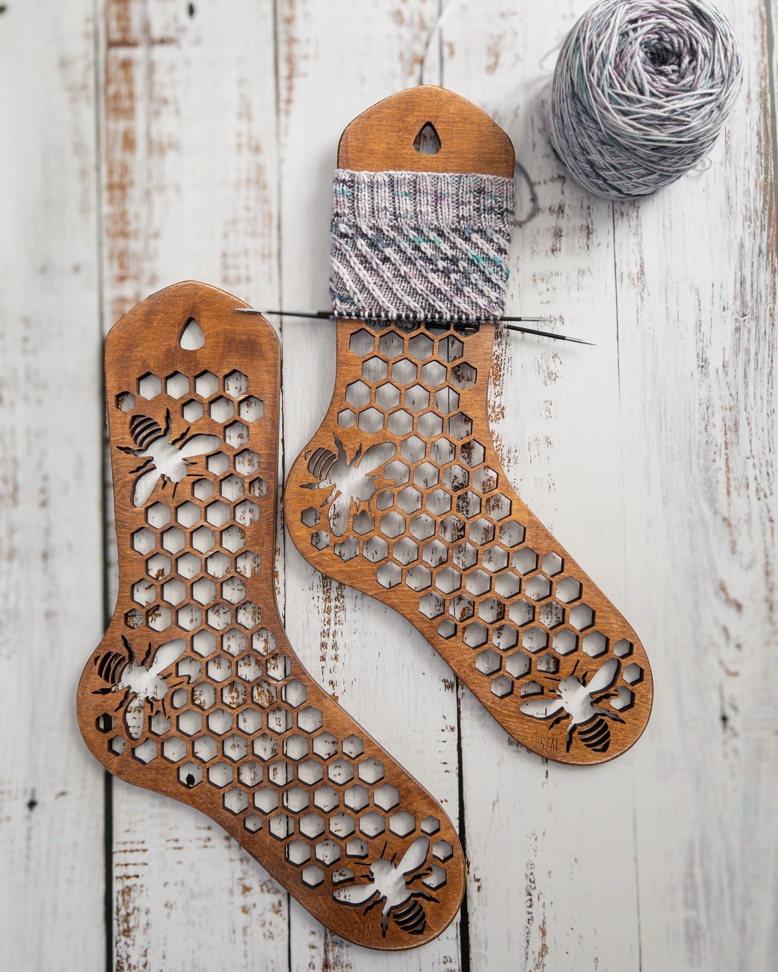 A pair of wood sock blockers with a honeycomb and bee motif. The start of a sock is on one of the blockers it’s made out of a gray variegated yarn and has diagonal striped stitched pattern. The ball of the yarn is nearby and the whole scene is on a white washed wooden surface .