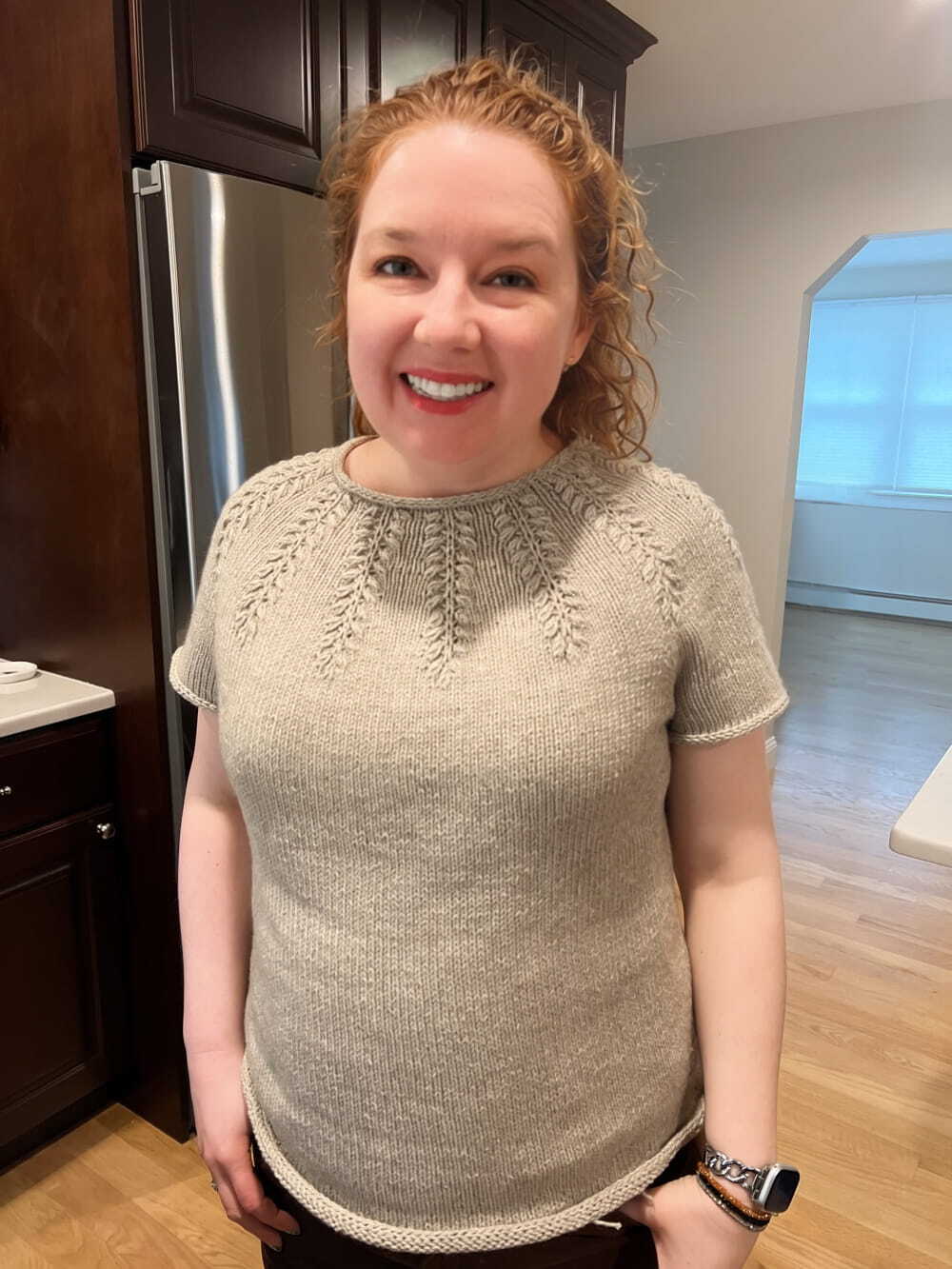 A beautiful and incredibly talented woman with curly red hair smiles while modeling a finished short-sleeve knitted tee in a light beige color. The sweater features a cabled yoke detail that looks almost like wheat stalks and a simple stockinette body with i-cord finishes at the neck, sleeve, and hems. I'm in my kitchen with wood floors and dark cabinets and really should find a nicer setting for my photoshoot, bit this will do for now. Pattern: Raunioiset Sweater by Wallebeck Wool. Yarn: Knit Picks Woolen Cotton.