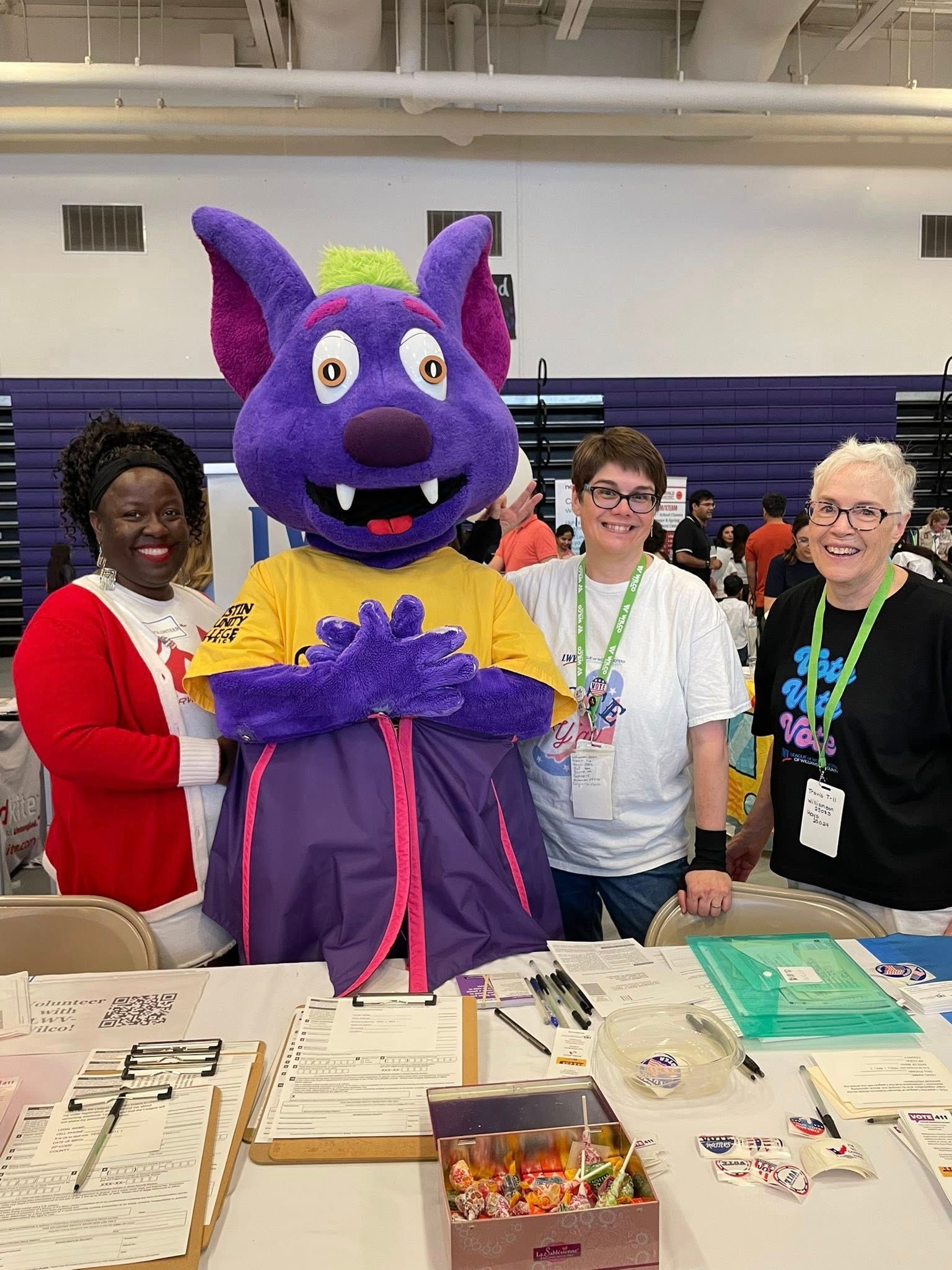 Celeste, Felicia, and Diane stand next to a purple mascot representing the ACC Riverbats. They're all behind a table filled with voter registration forms.