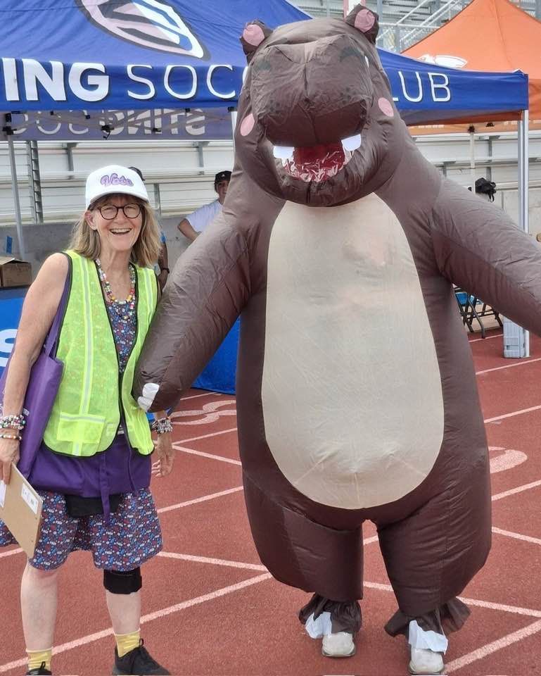 Helen wears a white hat, yellow hi-viz vest, and a blue dress with VOTE printed all over and stands next to a person wearing an inflatable hippo mascot costume at the Hutto back to school rally in July