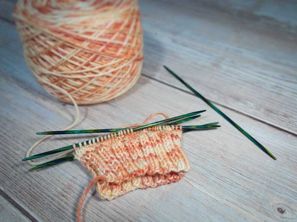 A small section of hand knitting in progress worked in variegated peach and cream yarn on green double-pointed needles. The fabric shows a textured stitch pattern and is partially folded. A matching ball of yarn sits nearby on a light wooden surface, with one needle set aside.