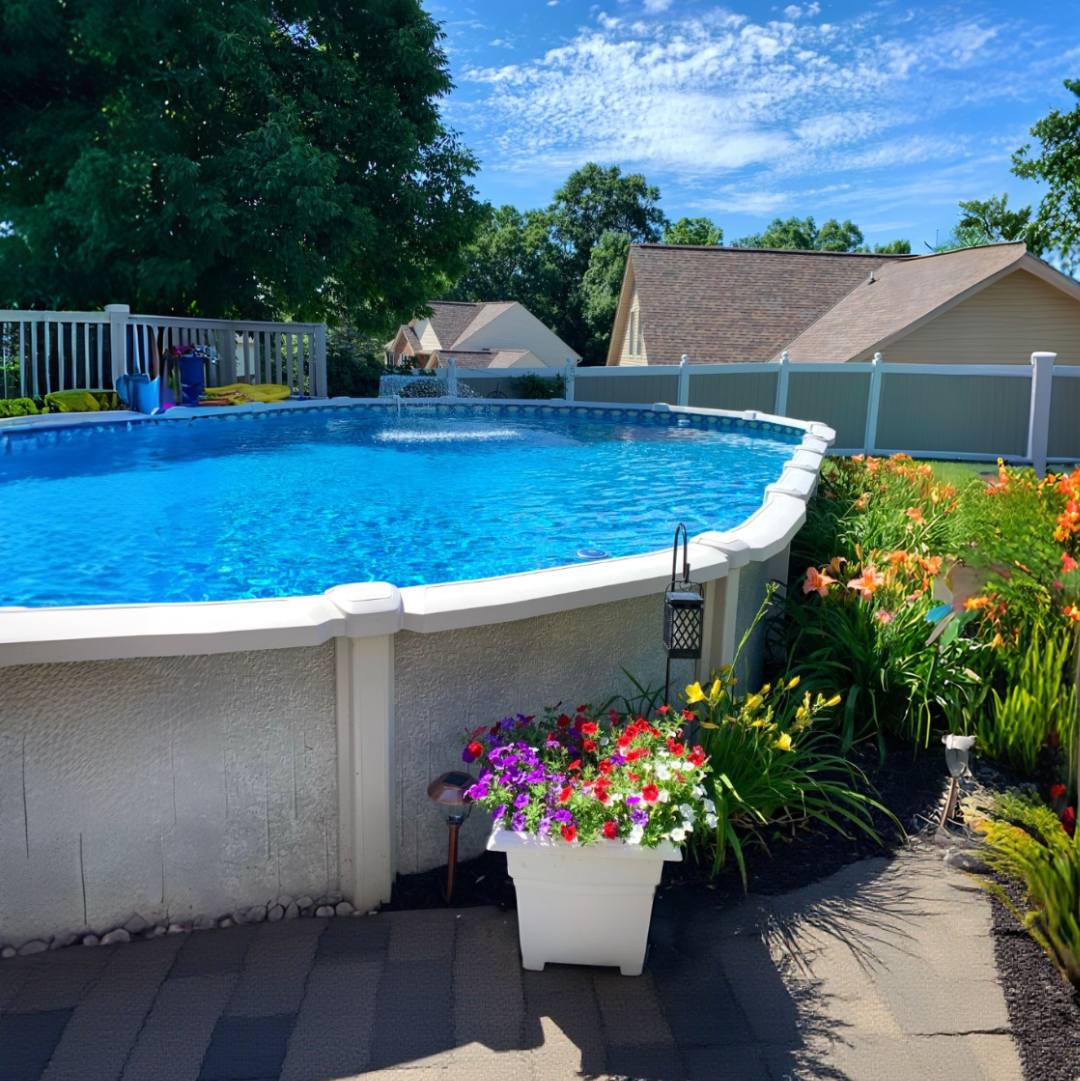 Sunny backyard featuring a round above ground swimming pool with blue water, colorful flowers and landscaping, patio pavers, white fence, and bright blue sky with clouds.