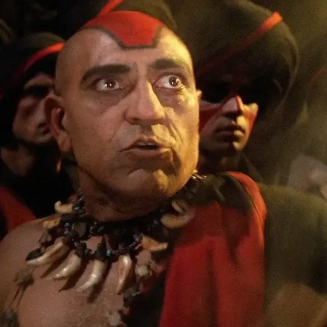 Gros plan d'un grand prêtre au crâne rasé, maquillage rouge sur le front et collier de griffes, entouré de gardes en tenue rouge et noire dans une scène inspirée d'Indiana Jones et le Temple maudit.